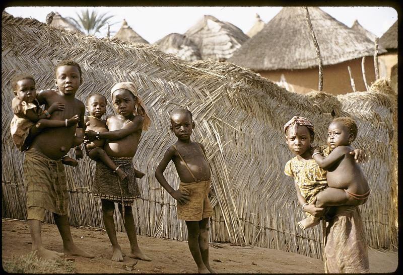 Children standing along a matting fence of cane and reed, with thatched compounds behind them.