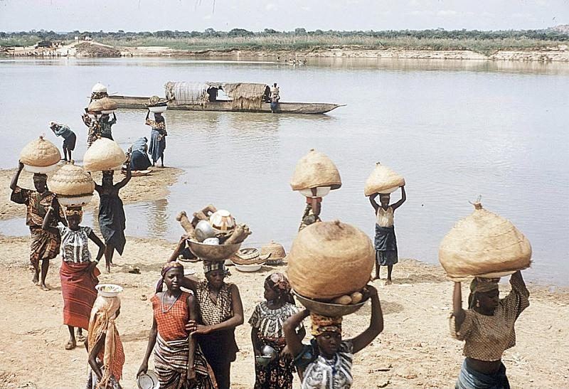 People carrying large lidded baskets and enamel basins up from a river landing on the Niger, with canoes behind them.
