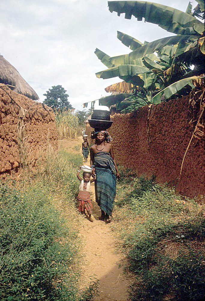 A woman carrying a load on her head walking a narrow path between high mud walls.