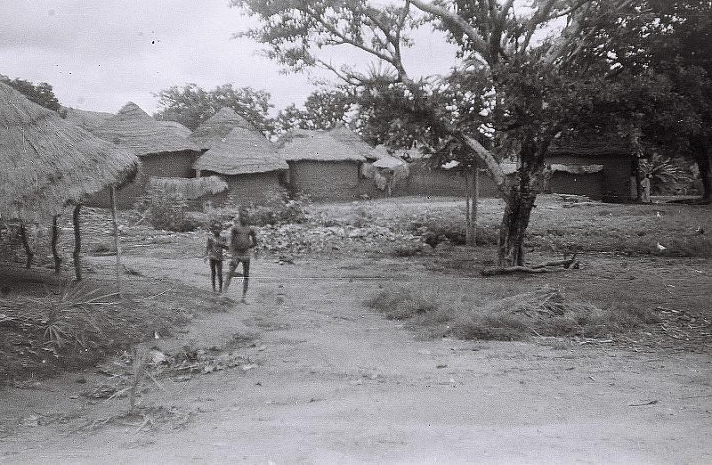 Thatched compounds and open ground photographed in black and white.