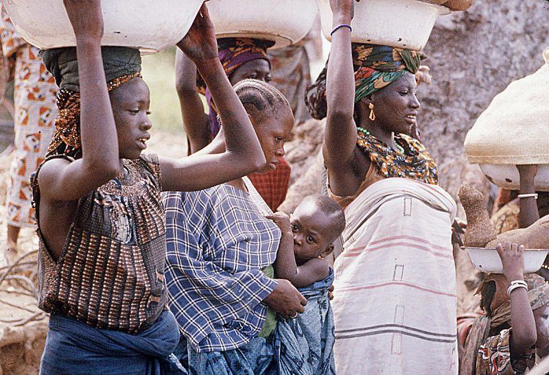 Women carrying enamel basins on their heads, in printed cotton, indigo and hand-woven striped cloth.